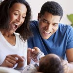 Happy mixed race young adult parents playing with their three month old baby son, lying on his motherâ»u20acs knee, selective focus