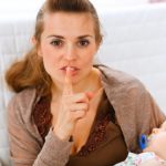 Young mama holding sleeping baby and showing shh gesture at home
