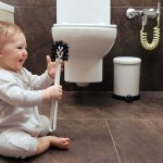 Happy toddler baby boy is playing in the toilet room with a brush. Smiling child plays on the brown floor in the beige bathroom