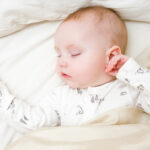 Infant with white bodysuit sleeping in parents bed in morning. Baby lying on back. Closeup. Top down view.
