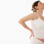 Pregnant woman holding back in front of white curtain