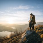 Pregnant mother and toddler stand on rock above lake at sunset