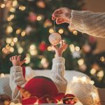 Hand of mother holding gold Christmas tree balls with playful baby enjoying near the Christmas tree in holiday, little infant girl in Santa Claus hat, First Christmas, Christmas and New Year concept.